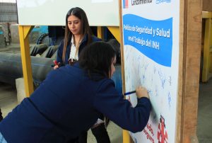 Funcionaria firma el compromiso con la Política de Seguridad del INH, con lápiz sobre un afiche de la política.